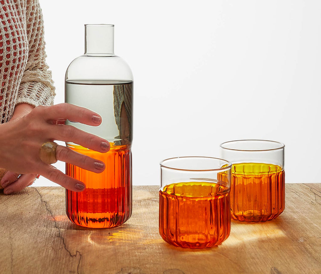 Person holding an amber and smoke carafe with matching glasses on a wooden surface.