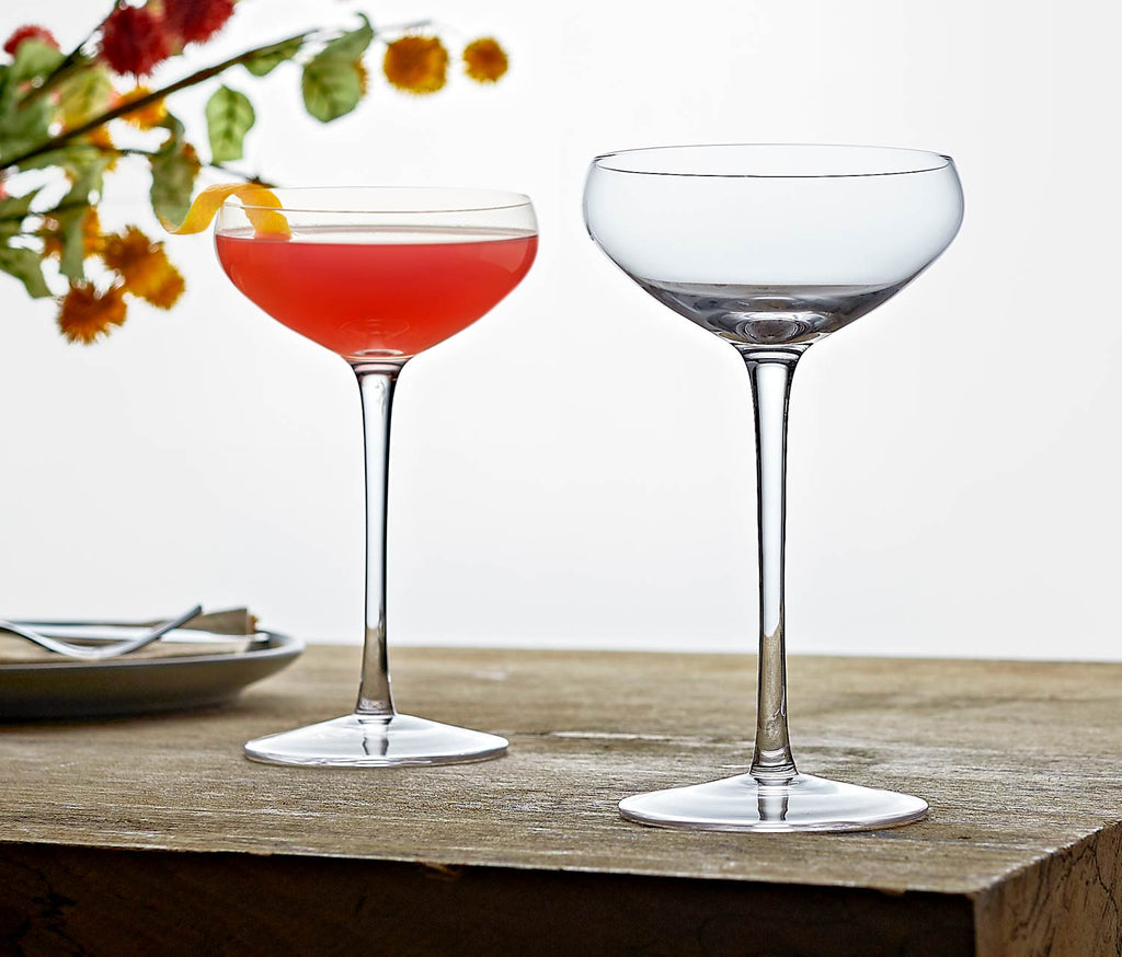 Two modern elegant cocktail glasses on a wooden surface with one containing a cocktail.
