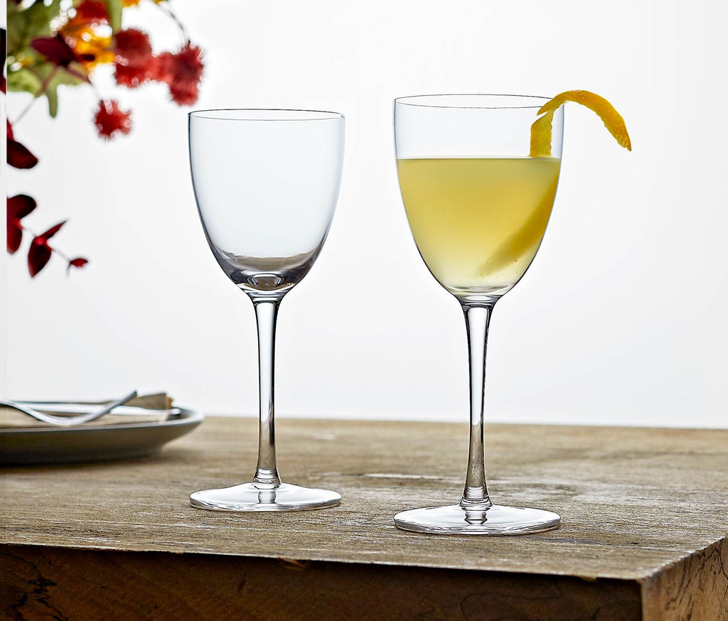 Two elegant Nick and Nora glasses on a wooden surface, with one containing a martini cocktail and a lemon twist.
