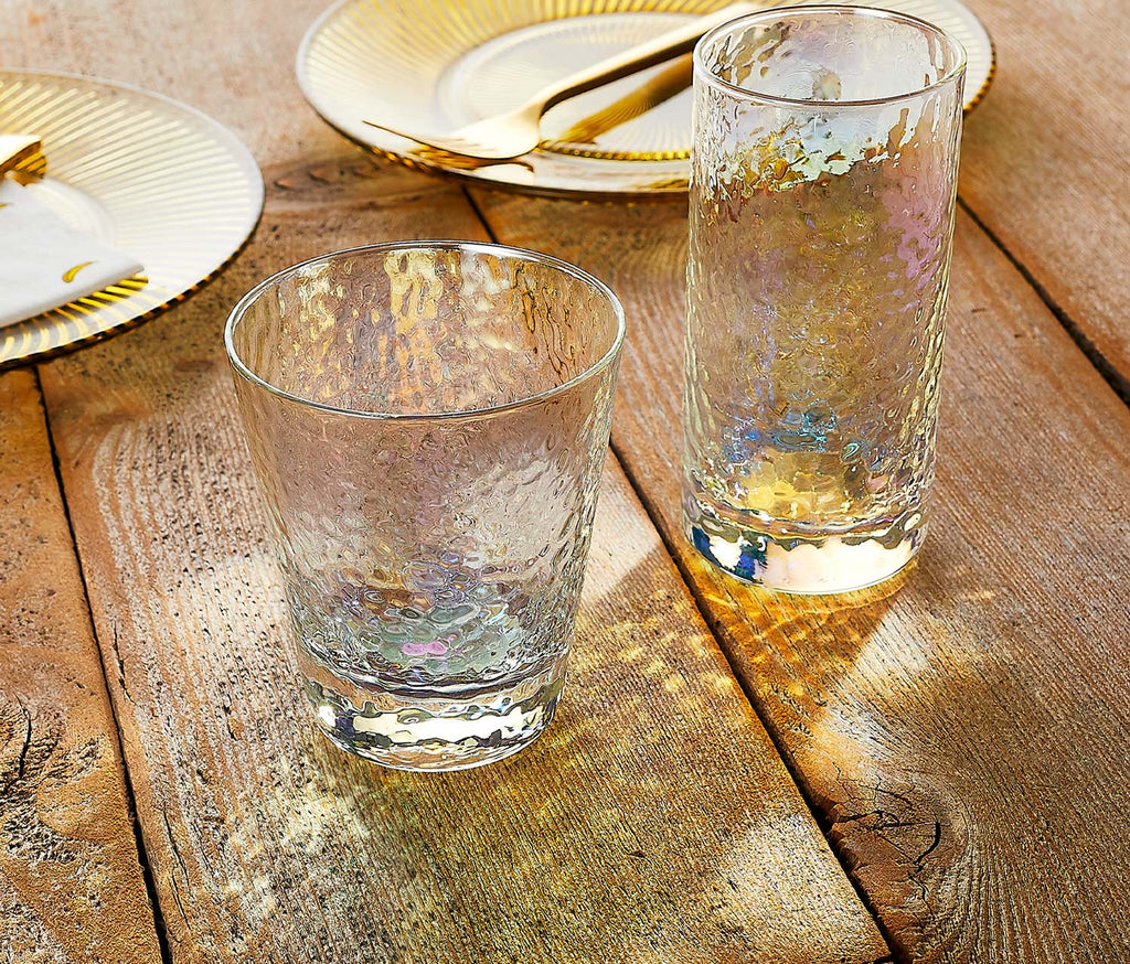 Hammered rainbow iridescent glass tumblers on a wooden surface with gold-rimmed plates in the background.