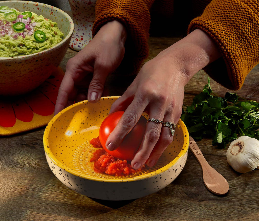 Person preparing a dish with a grate plate of guacamole and a tomato on a wooden surface.