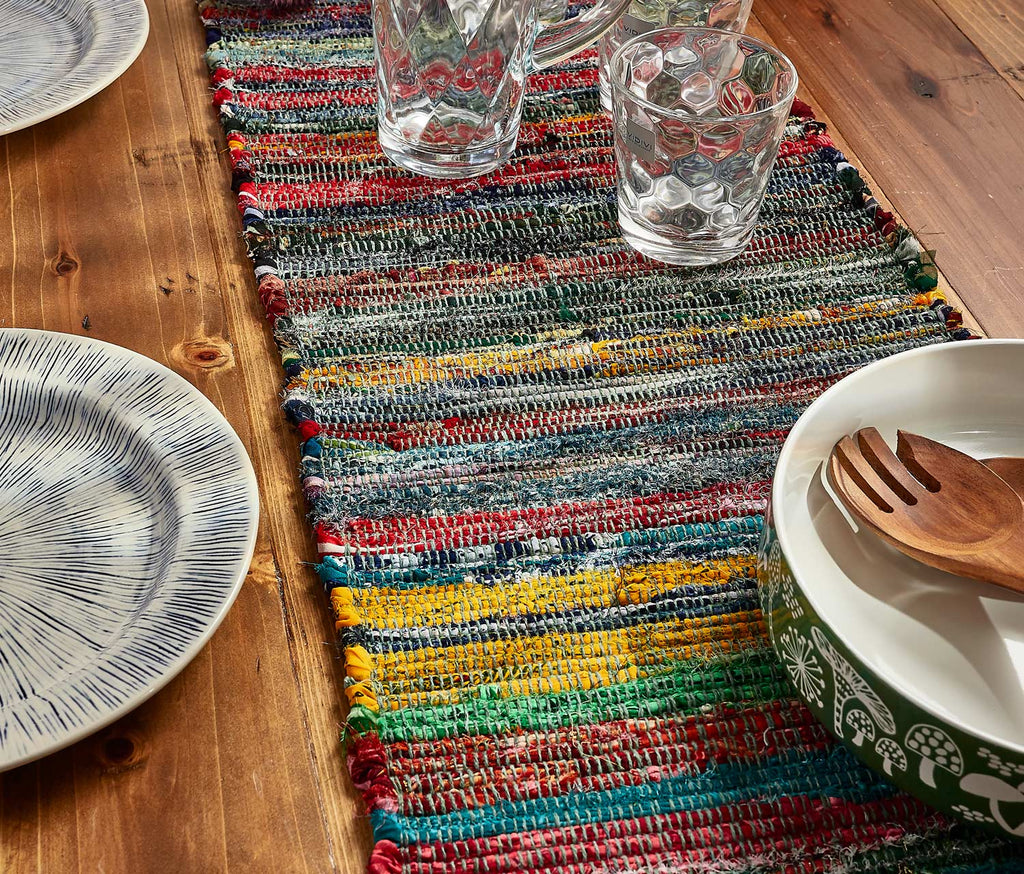 Colorful woven table runner on a wooden table with plates, glasses, and a bowl.