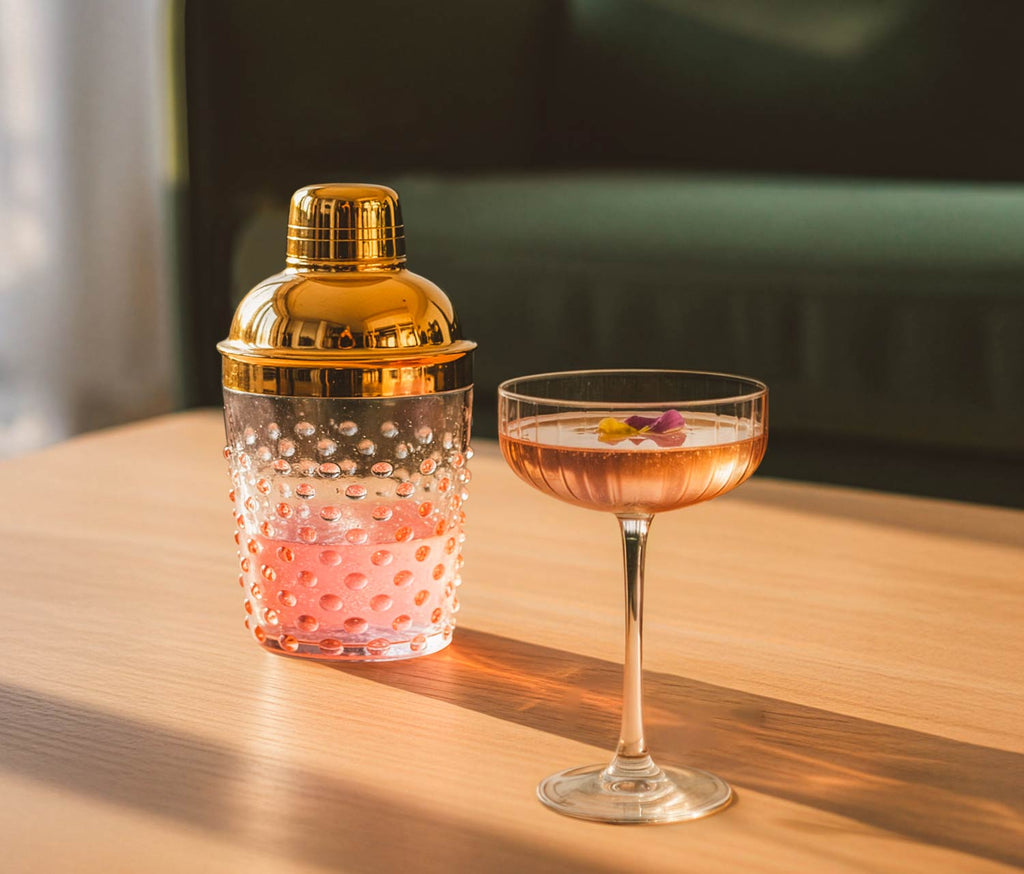 Gold cocktail shaker and pink coupe with pink drink on a wooden table -Lollygag