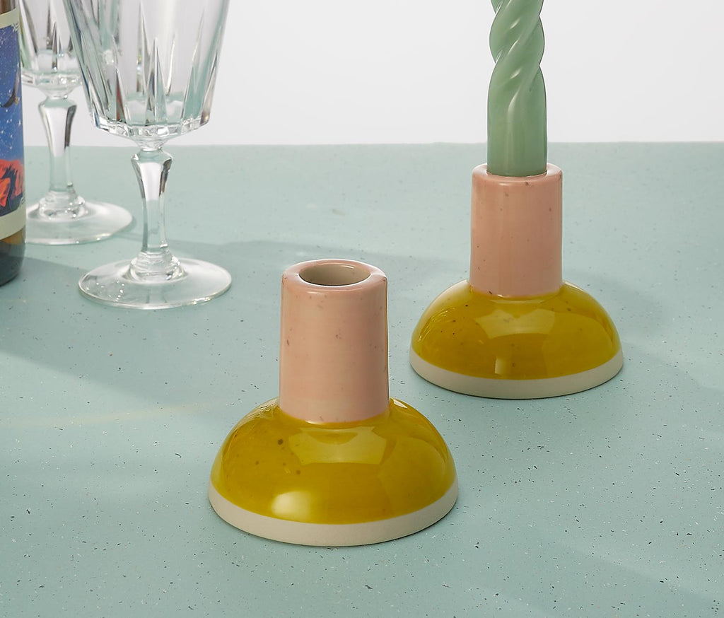 Two ceramic candle holders on a light blue surface with glasses and a bottle in the background.