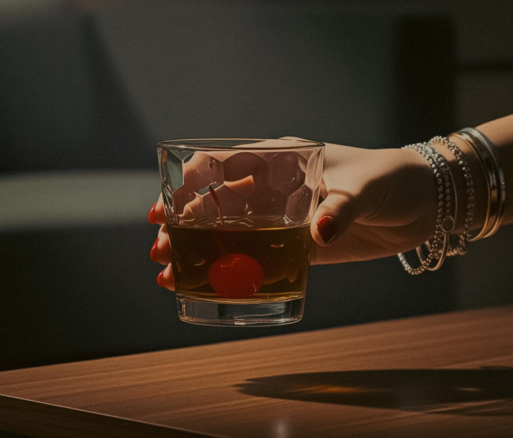 a woman's Hand holding a honeycomb glass of whiskey with cherries against a dark background