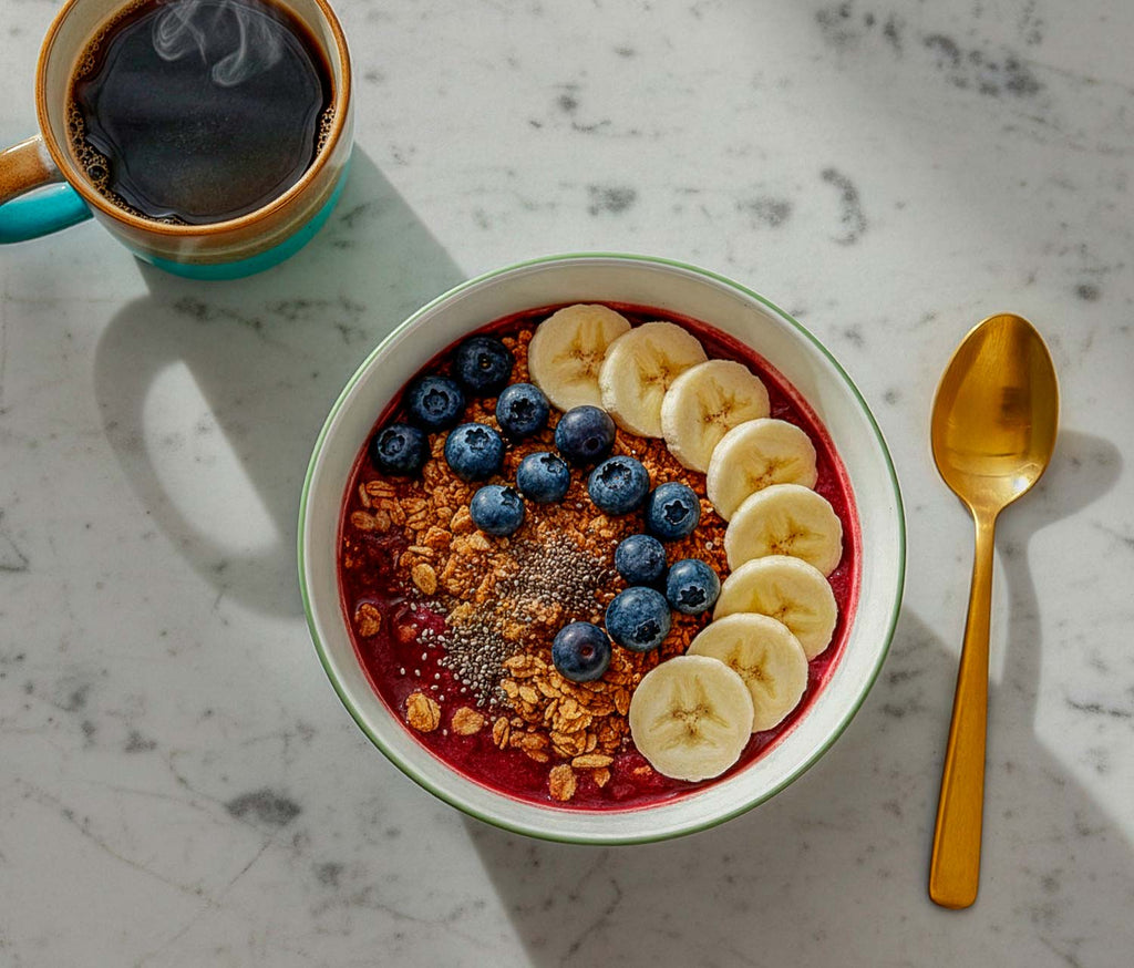 A bowl of acai bowl with bananas and blueberries, accompanied by a cup of coffee on a marble surface.